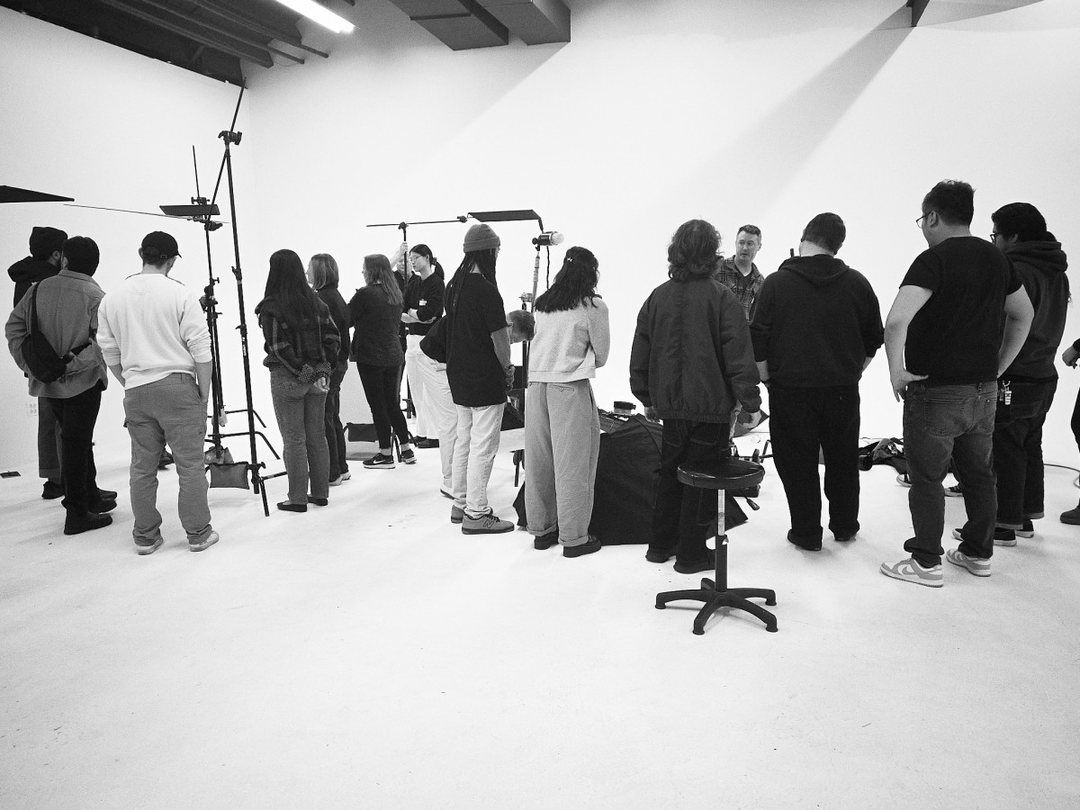 Students gather around light stands and flags in a white studio as a photographer in plaid explains the setup.
